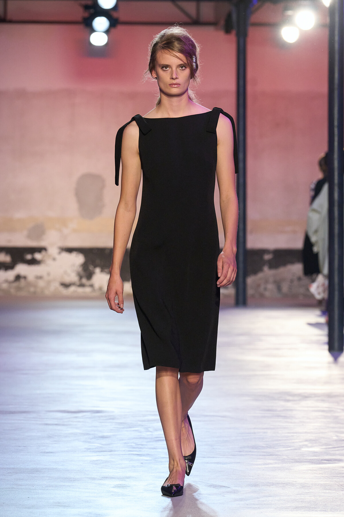 Model walks the runway in a knee-length black sheath dress with tied shoulder straps.