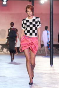 Model walks runway in a black-and-white checker knit top with a pink satin draped mini skirt, pink lighting in the background.