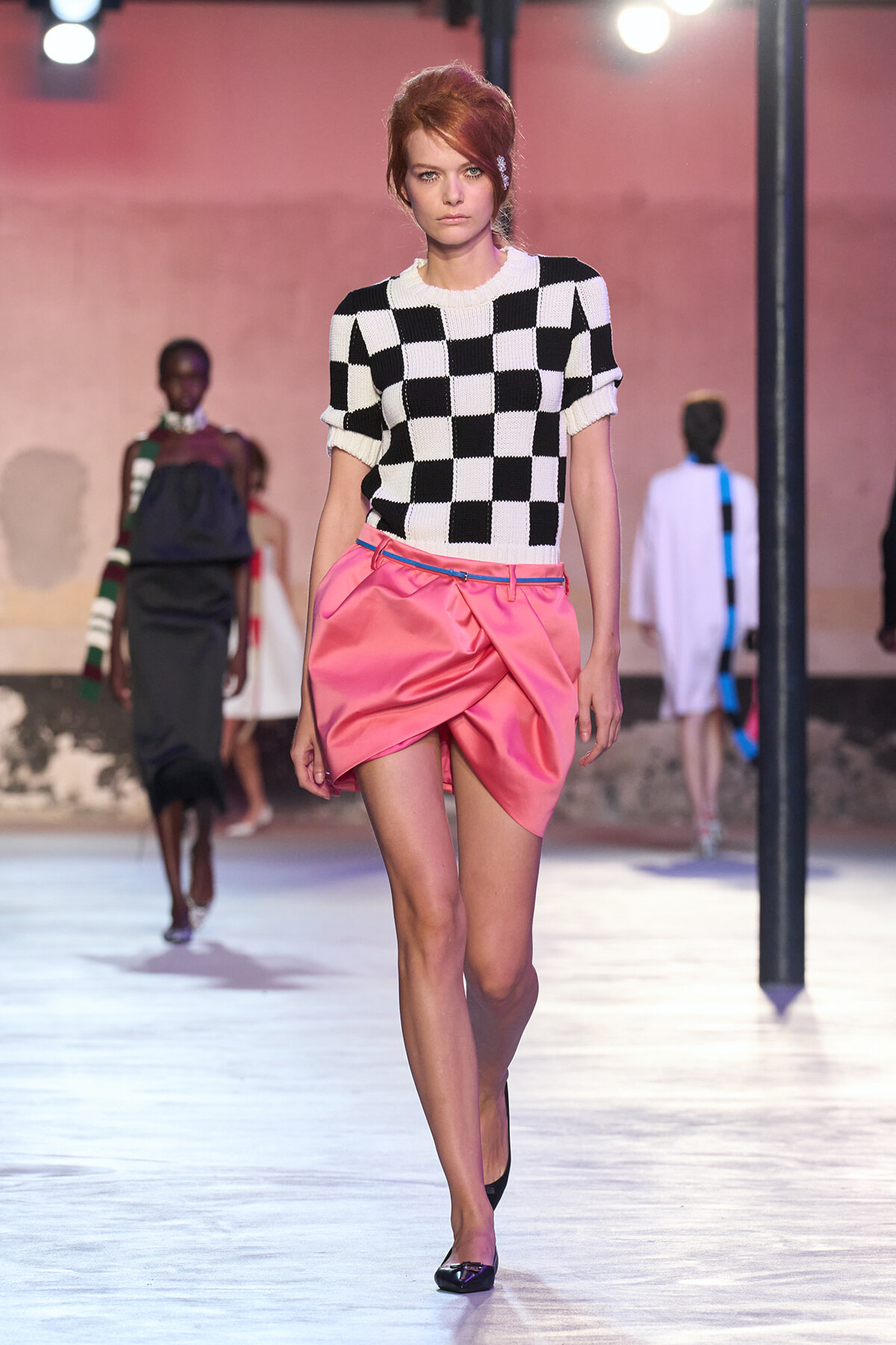 Model walks runway in a black-and-white checker knit top with a pink satin draped mini skirt, pink lighting in the background.