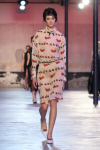 Model walking runway in a pale pink cherry-print dress with a pearl statement necklace and white heels.