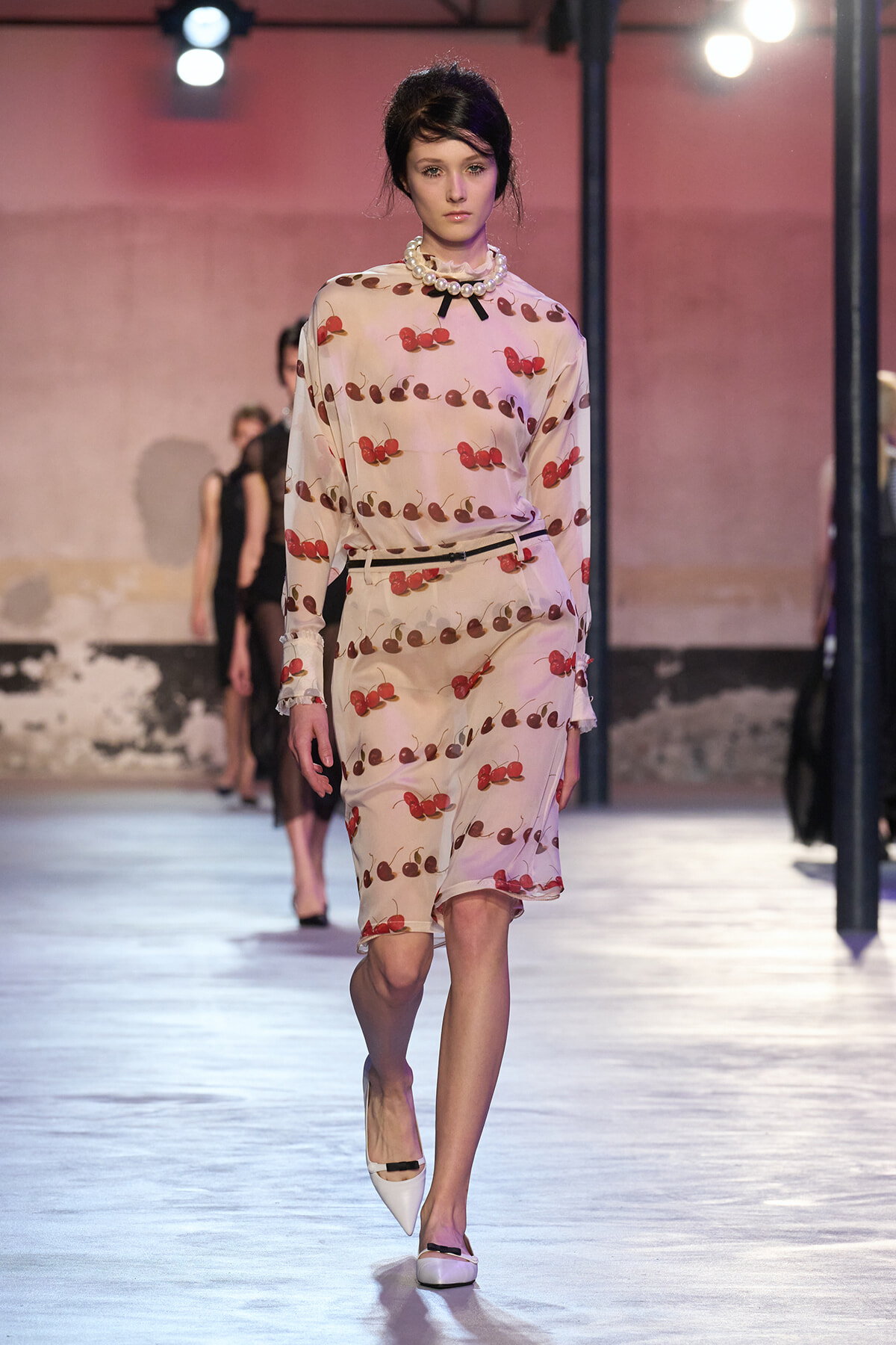 Model walking runway in a pale pink cherry-print dress with a pearl statement necklace and white heels.