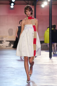 Model walks a fashion show wearing a white strapless knee-length dress with a long beige-red striped scarf draped over her neck and shoulder, on a bright runway.