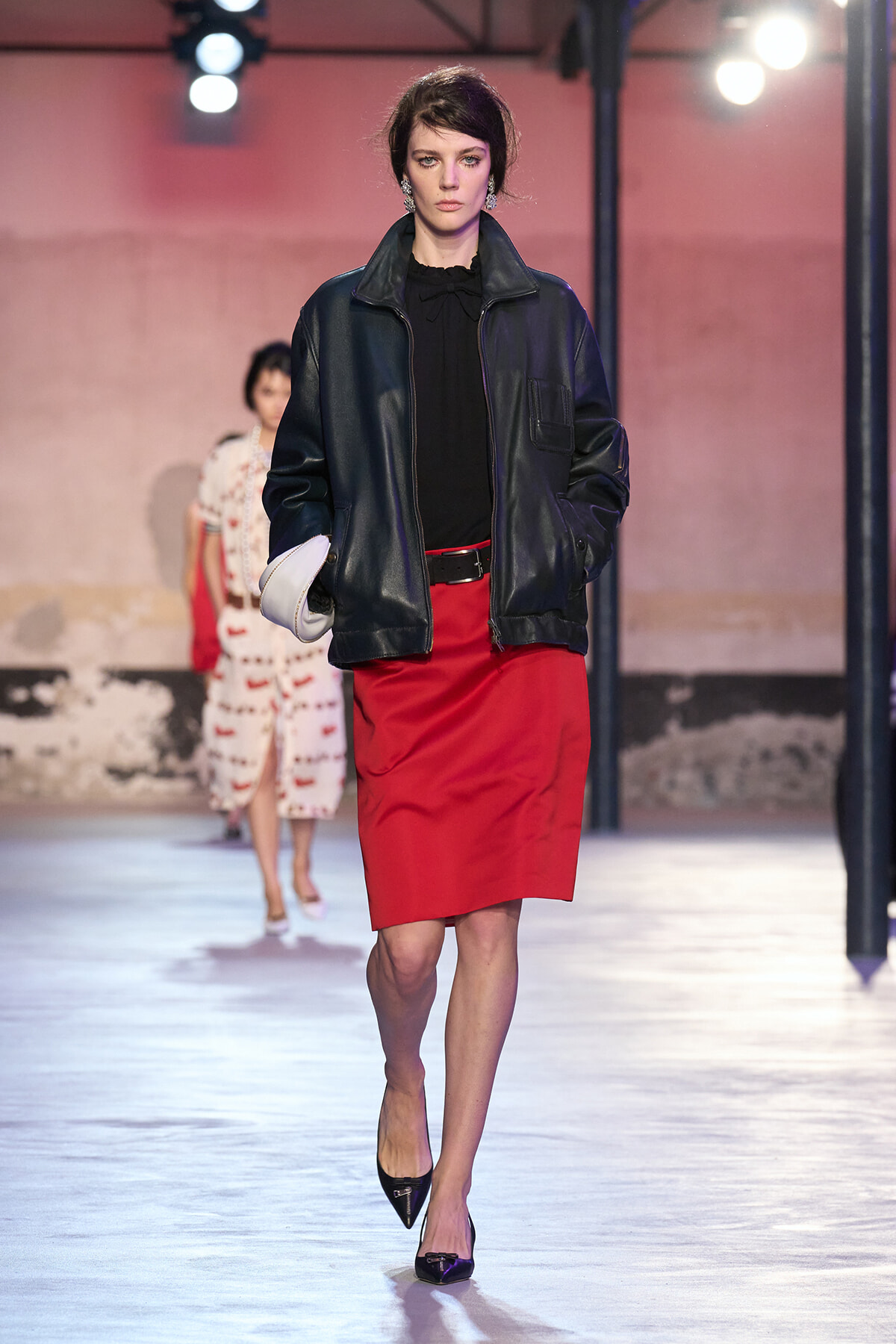Model walking the runway in a black leather jacket, black blouse, and red pencil skirt, carrying a white clutch; bright fashion-show lights in background.