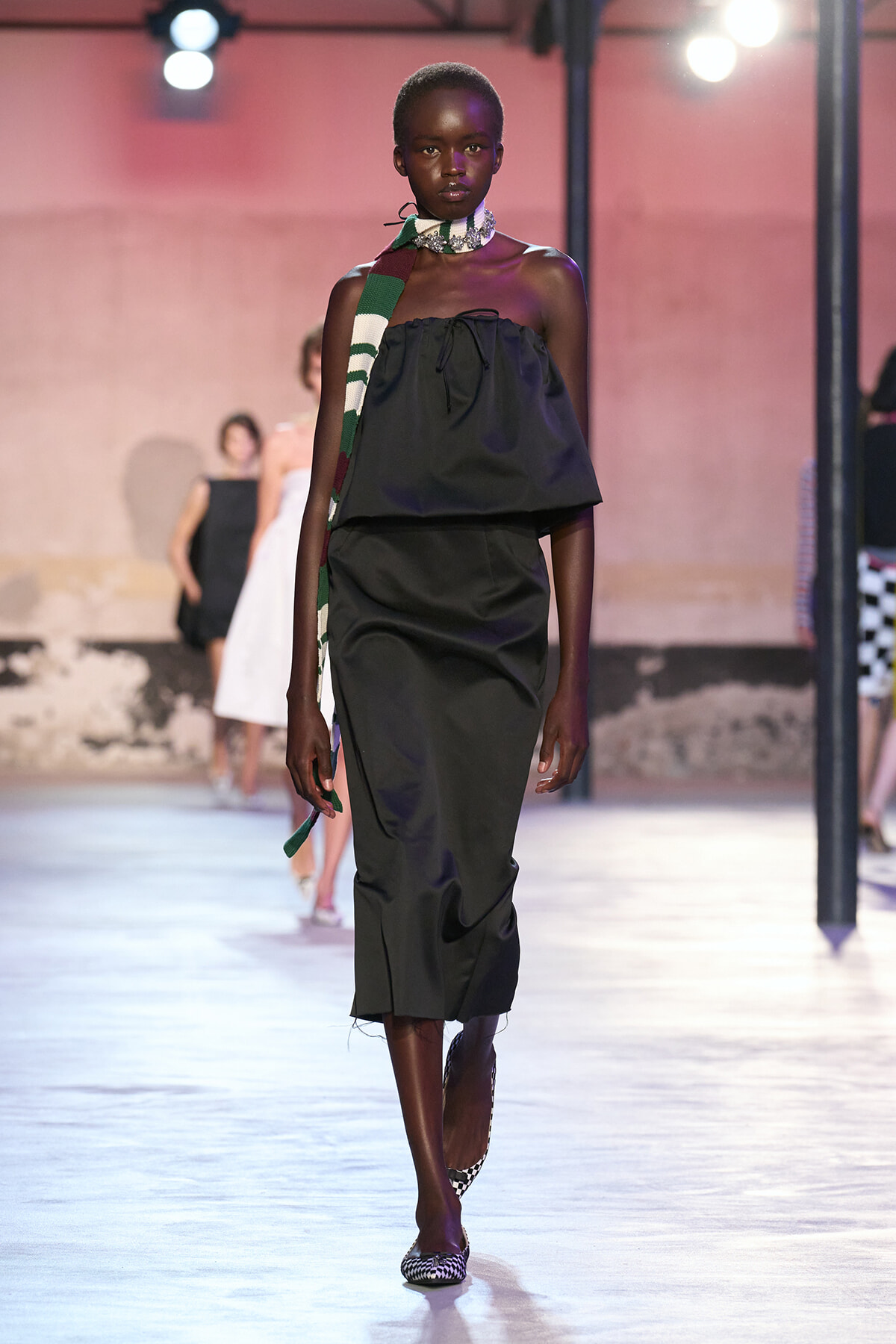 Model walking a runway in a strapless black top with a long dark skirt, accented by a green-and-white striped scarf and checkered flats.