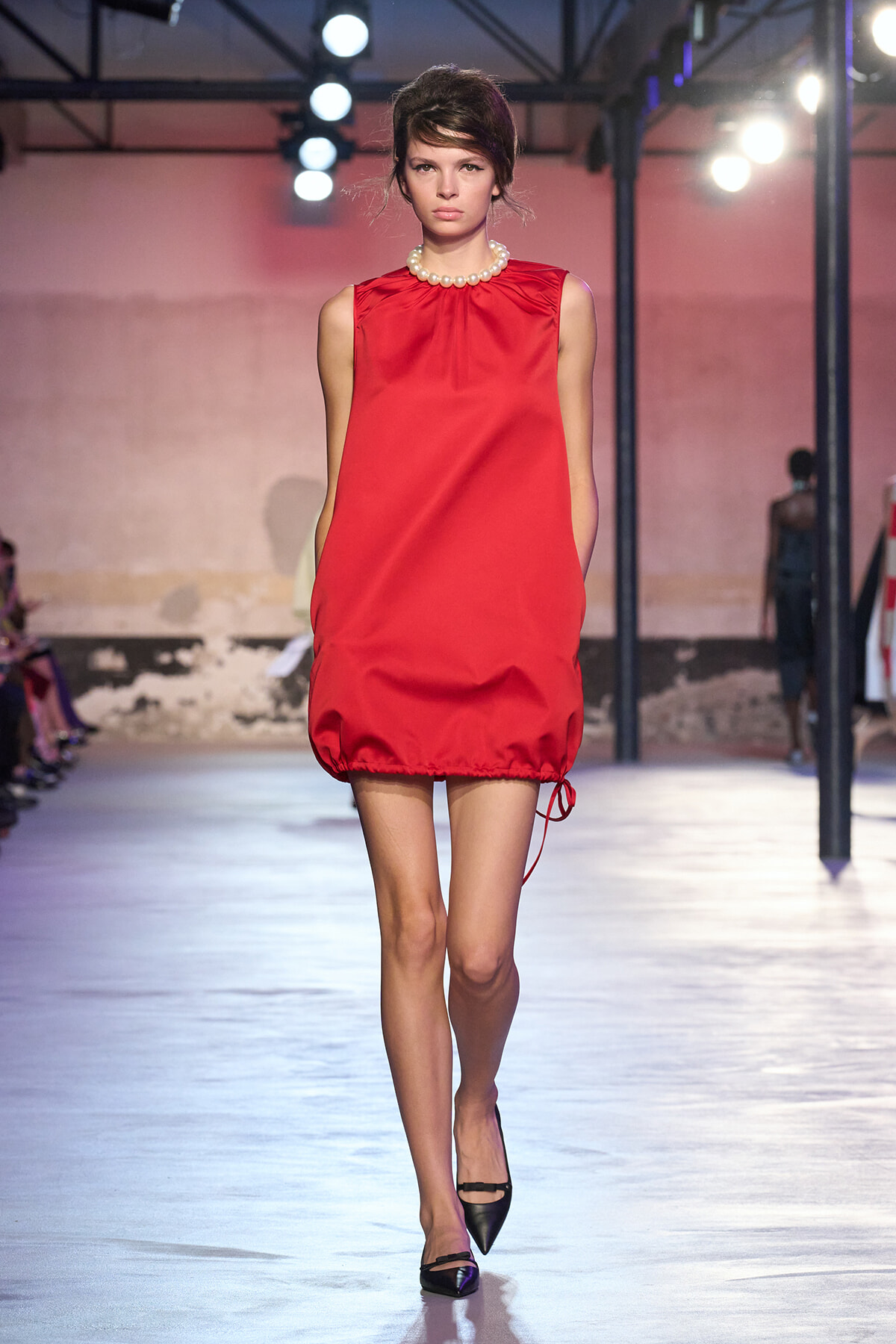 Model walking a runway in a bright red sleeveless shift dress with a pearl necklace and black heels.