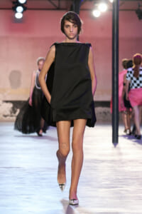 Runway model walking in a short black sleeveless dress with cape-like sides, white flats, and a small cat tattoo on her left thigh.