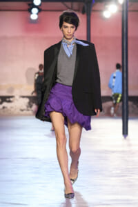Model struts a fashion runway in a black blazer, grey sweater vest, blue shirt with scarf, and a purple ruched mini skirt with checkered heels.