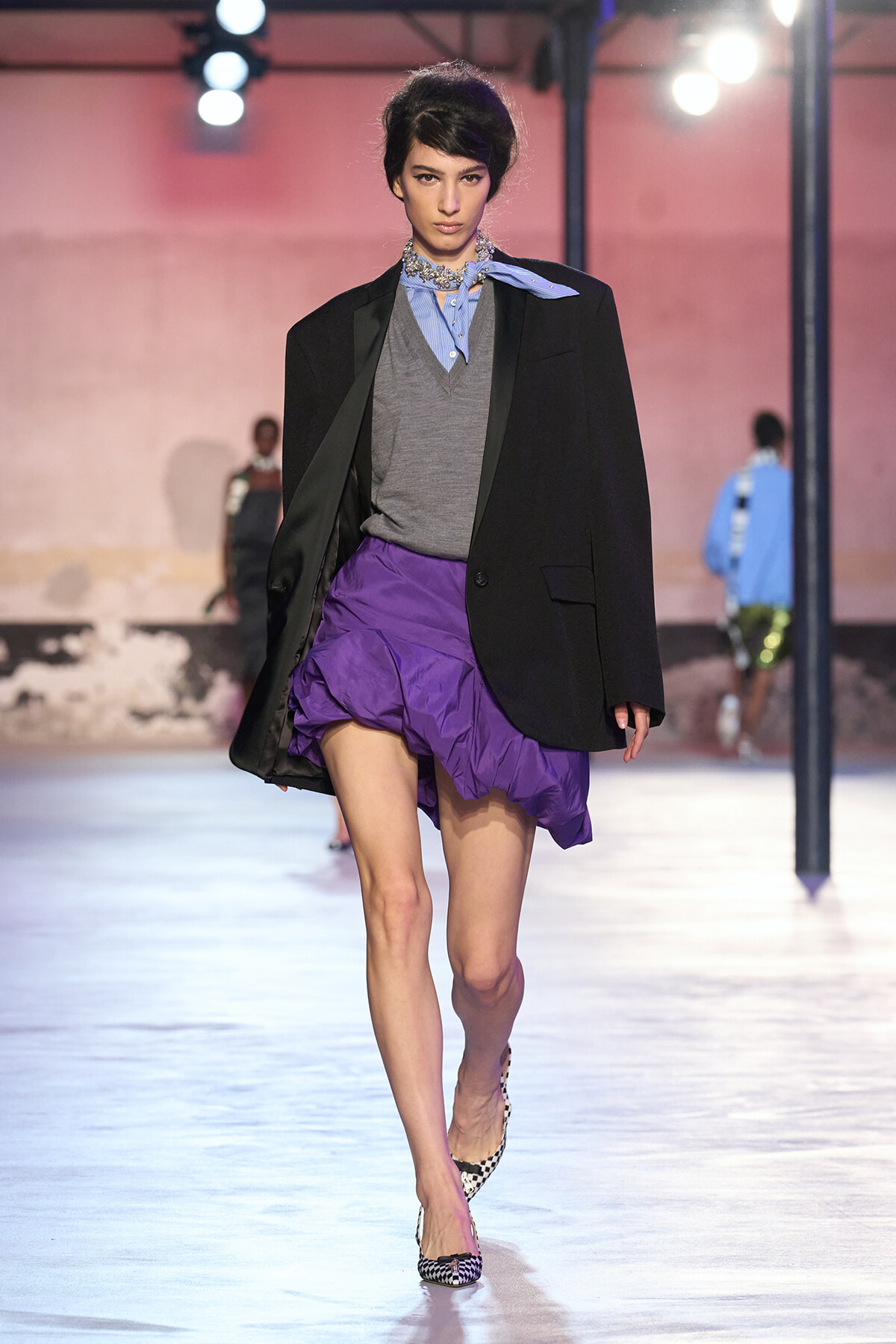 Model struts a fashion runway in a black blazer, grey sweater vest, blue shirt with scarf, and a purple ruched mini skirt with checkered heels.