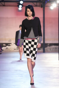 Model walking a fashion runway in a black cropped jacket over gray top with a pink hem, a pearl necklace, and a black-and-white checkerboard knit skirt, heels visible.