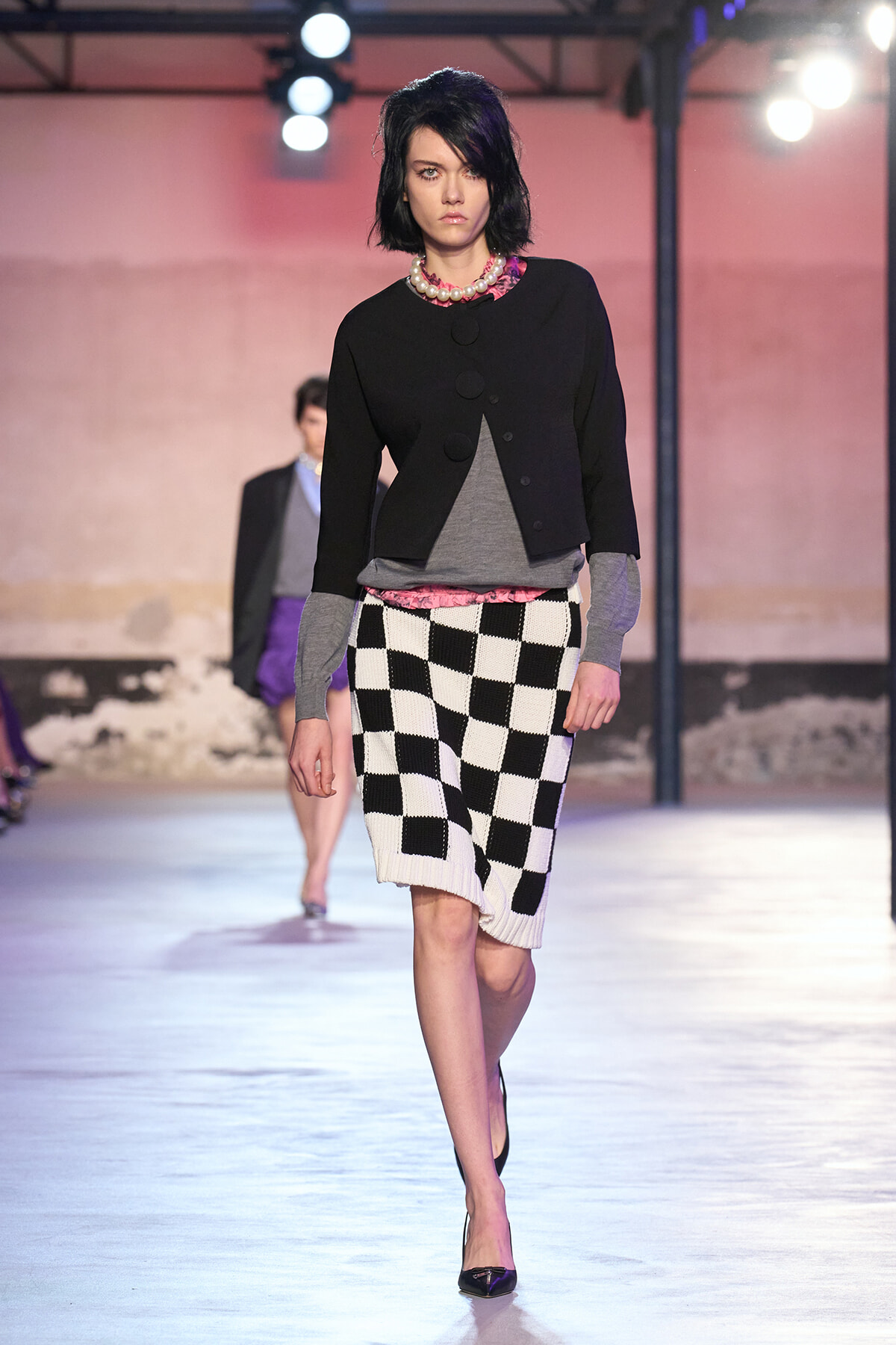 Model walking a fashion runway in a black cropped jacket over gray top with a pink hem, a pearl necklace, and a black-and-white checkerboard knit skirt, heels visible.