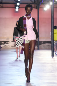 Runway model wearing a pink shirt dress with a black blazer and a purple striped scarf under bright stage lights