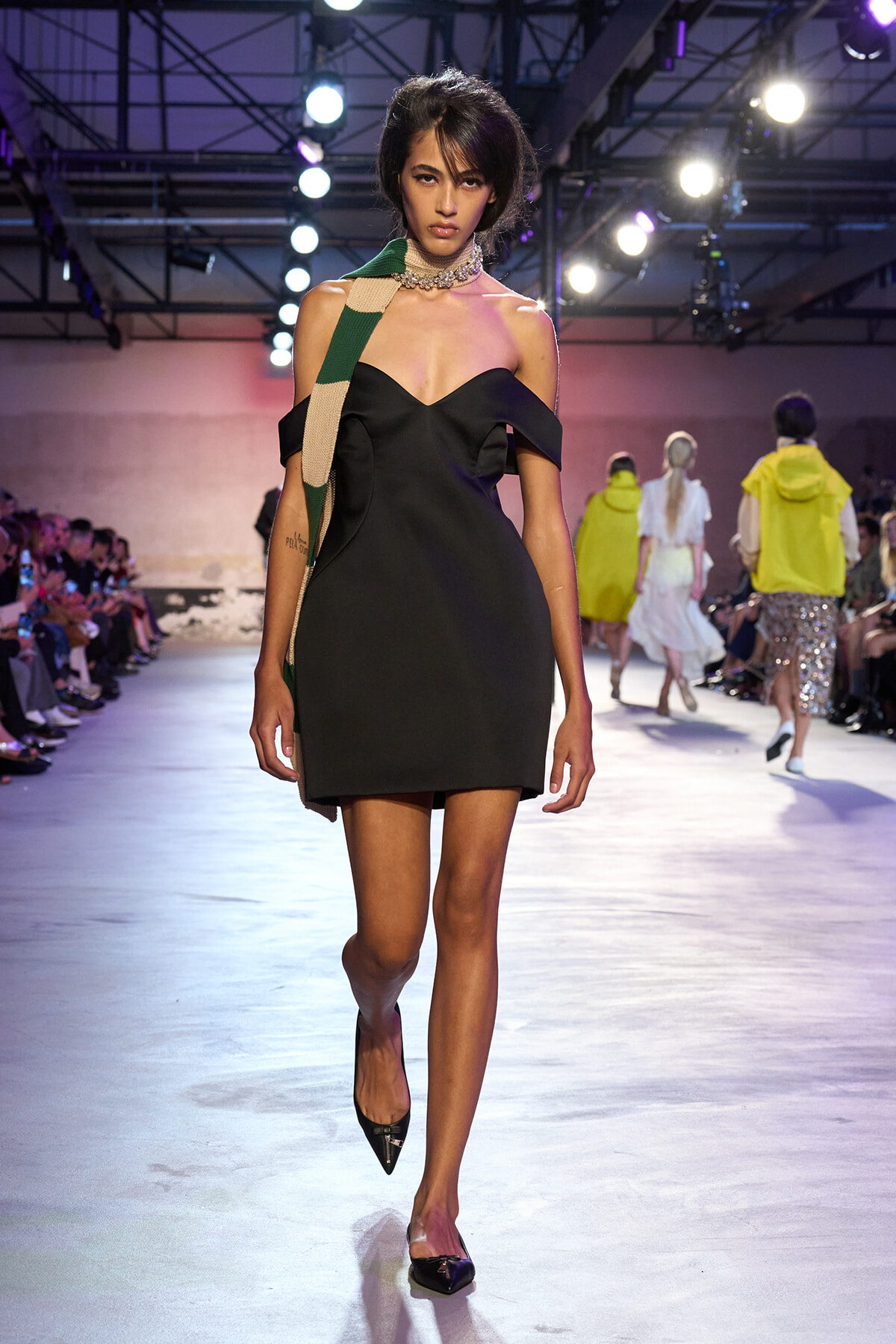Model walks a fashion runway in a black off-shoulder mini dress with a green-beige scarf and jeweled choker under bright stage lights.