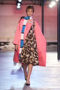 Model on runway wearing a pink coat over a sequined floral dress, paired with a blue-and-white striped scarf and black heels.