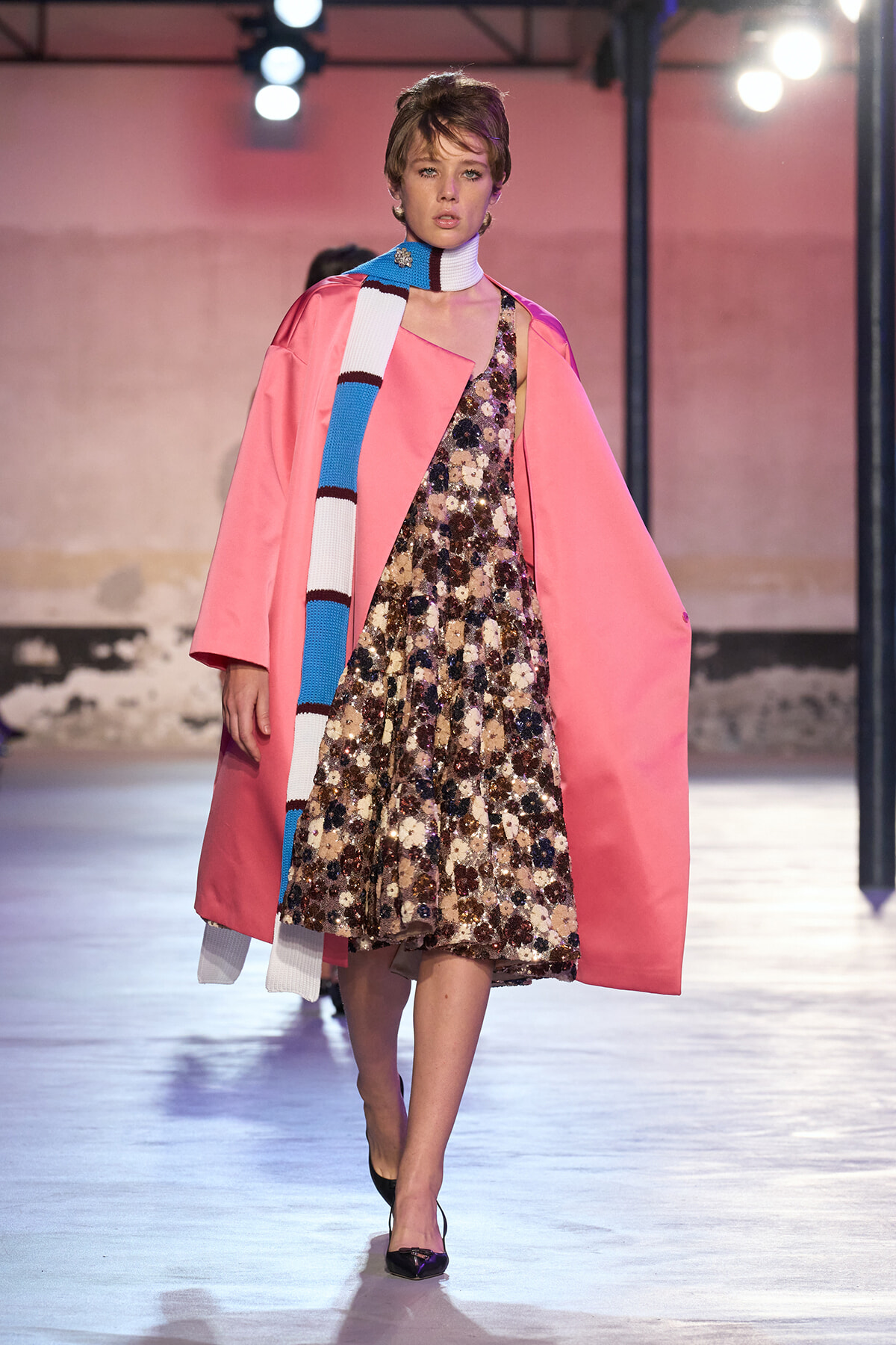 Model on runway wearing a pink coat over a sequined floral dress, paired with a blue-and-white striped scarf and black heels.