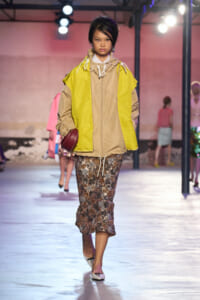 Model walks runway wearing a bright yellow and beige windbreaker over a beige jacket, paired with a sequined midi skirt and white flats, carrying a burgundy clutch.