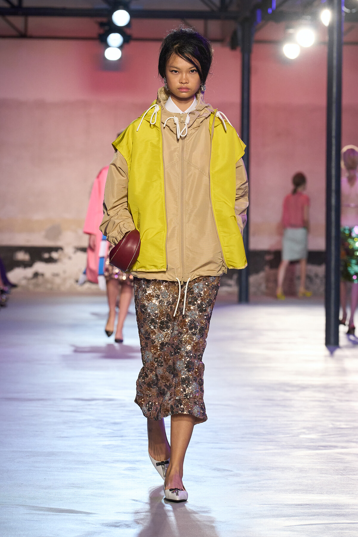 Model walks runway wearing a bright yellow and beige windbreaker over a beige jacket, paired with a sequined midi skirt and white flats, carrying a burgundy clutch.