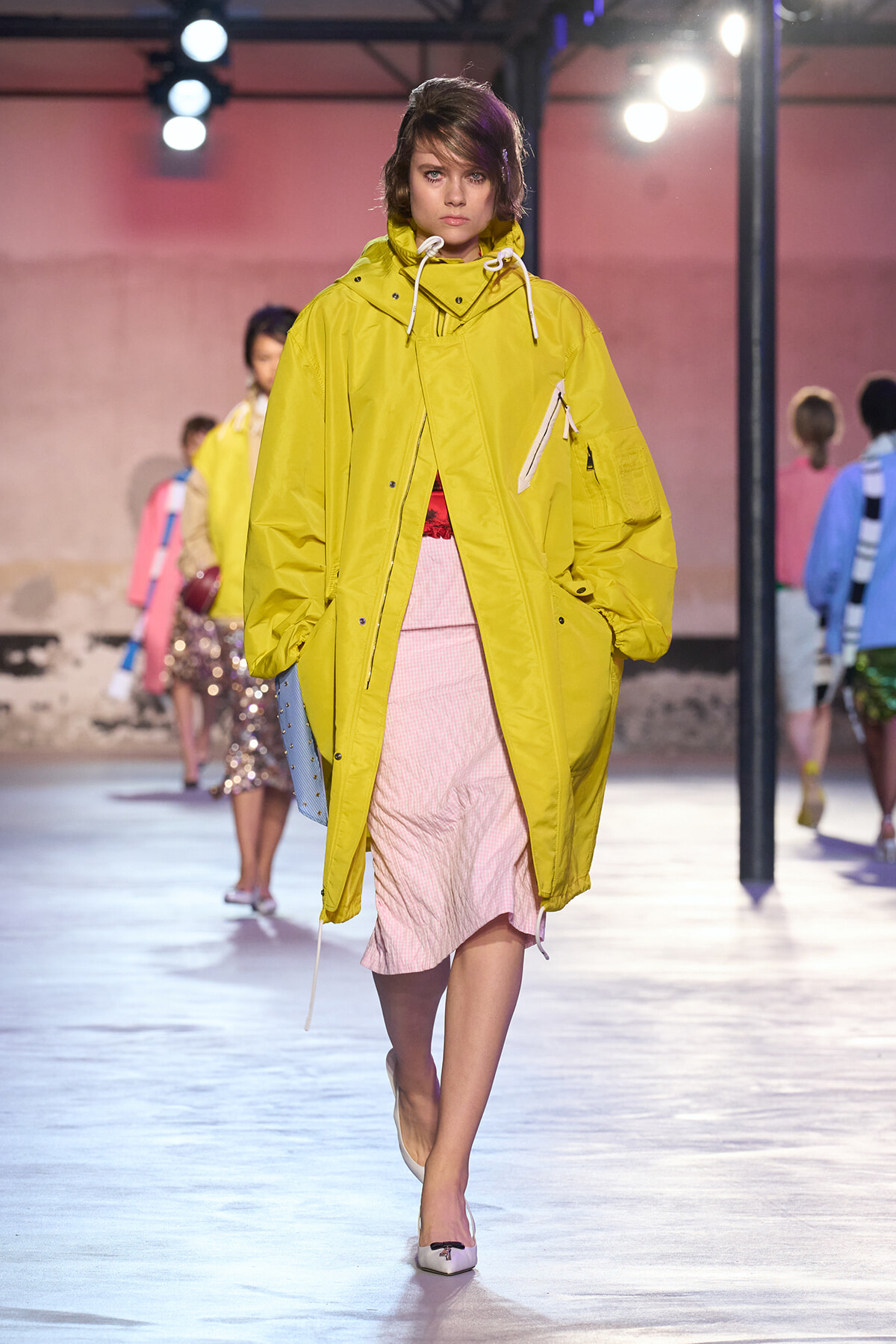 Model walking the runway in a bright yellow oversized parka over a pink dress, during a fashion show.
