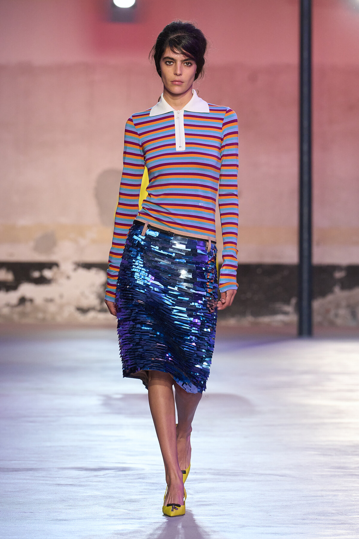 Model wearing a multicolor striped long-sleeve top with white collar and a blue sequin midi skirt on a runway.