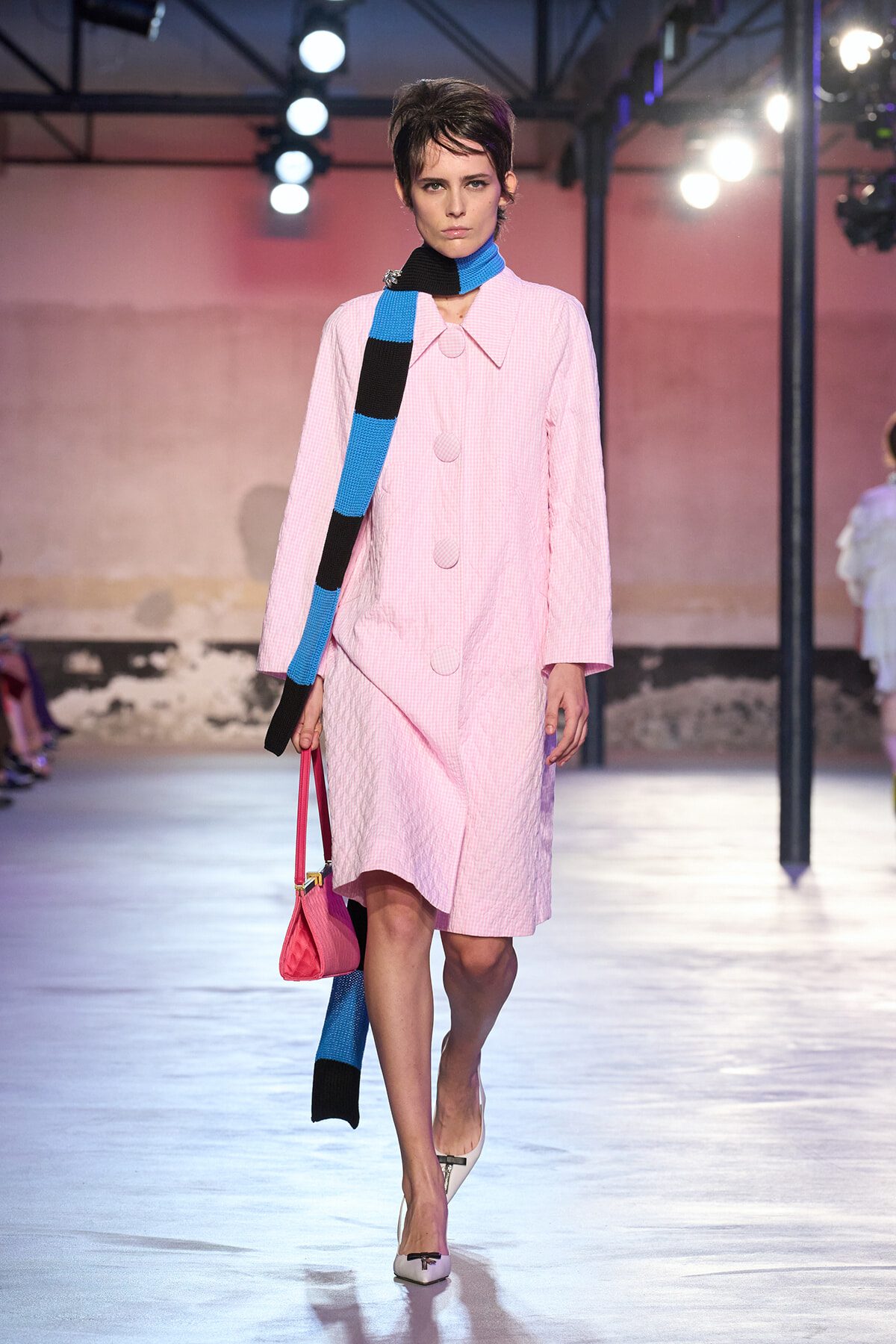 Runway model in a pink checkered coat with a blue-and-black striped scarf, carrying a pink bag and wearing white heels.