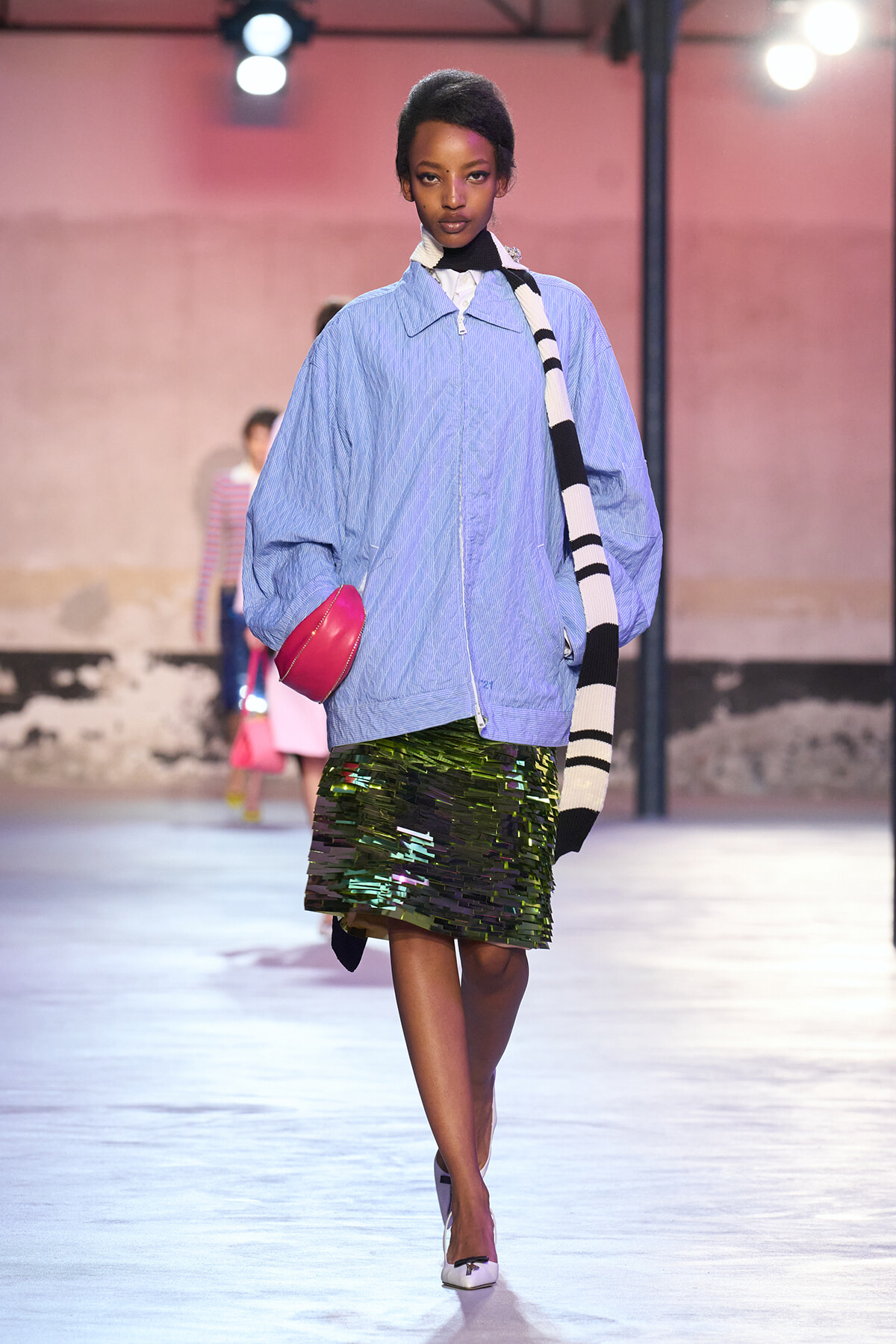 Model on a runway wearing a loose light-blue zip-up jacket, a shiny green sequin skirt, and a black-and-white striped scarf, holding a bright pink clutch.