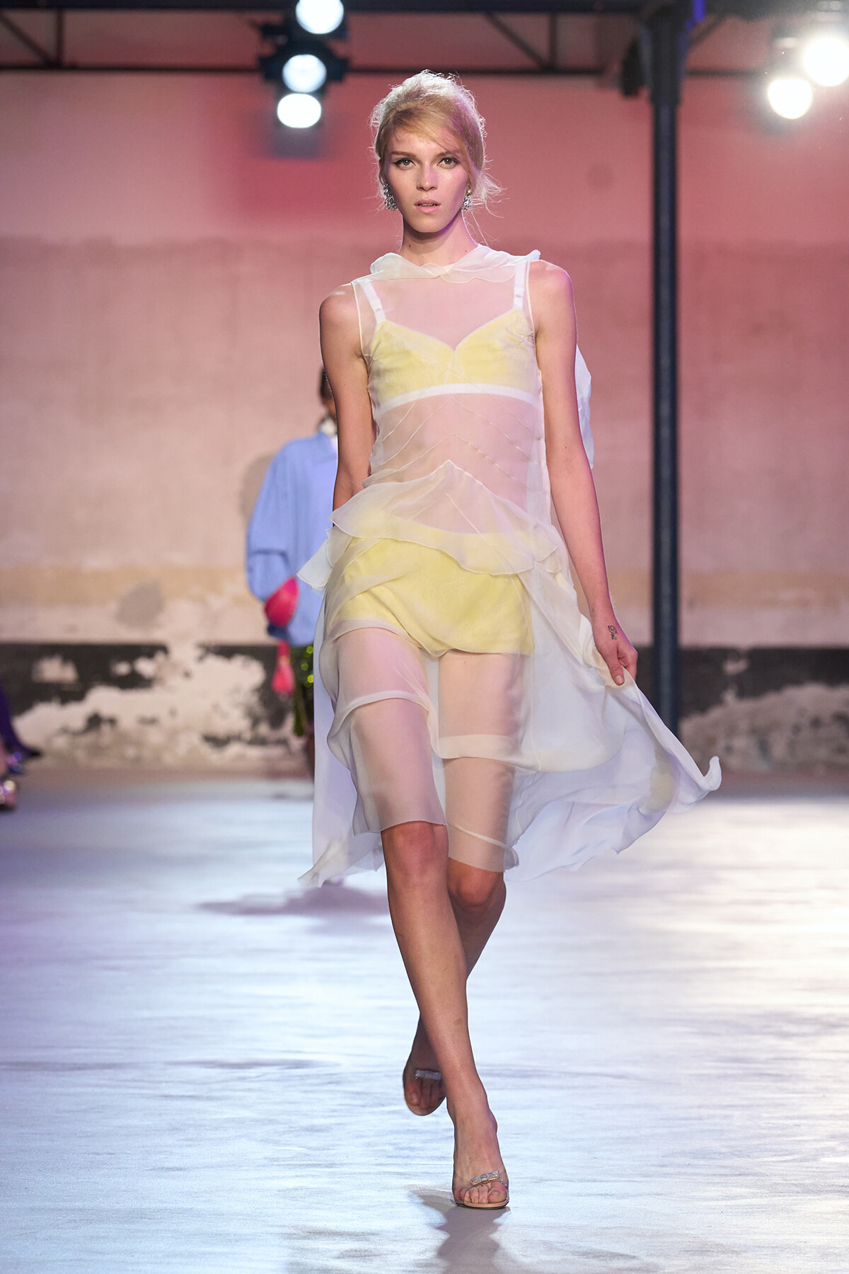 Model on a runway wearing a sheer, layered white dress with pale yellow undergarments, under bright show lights.