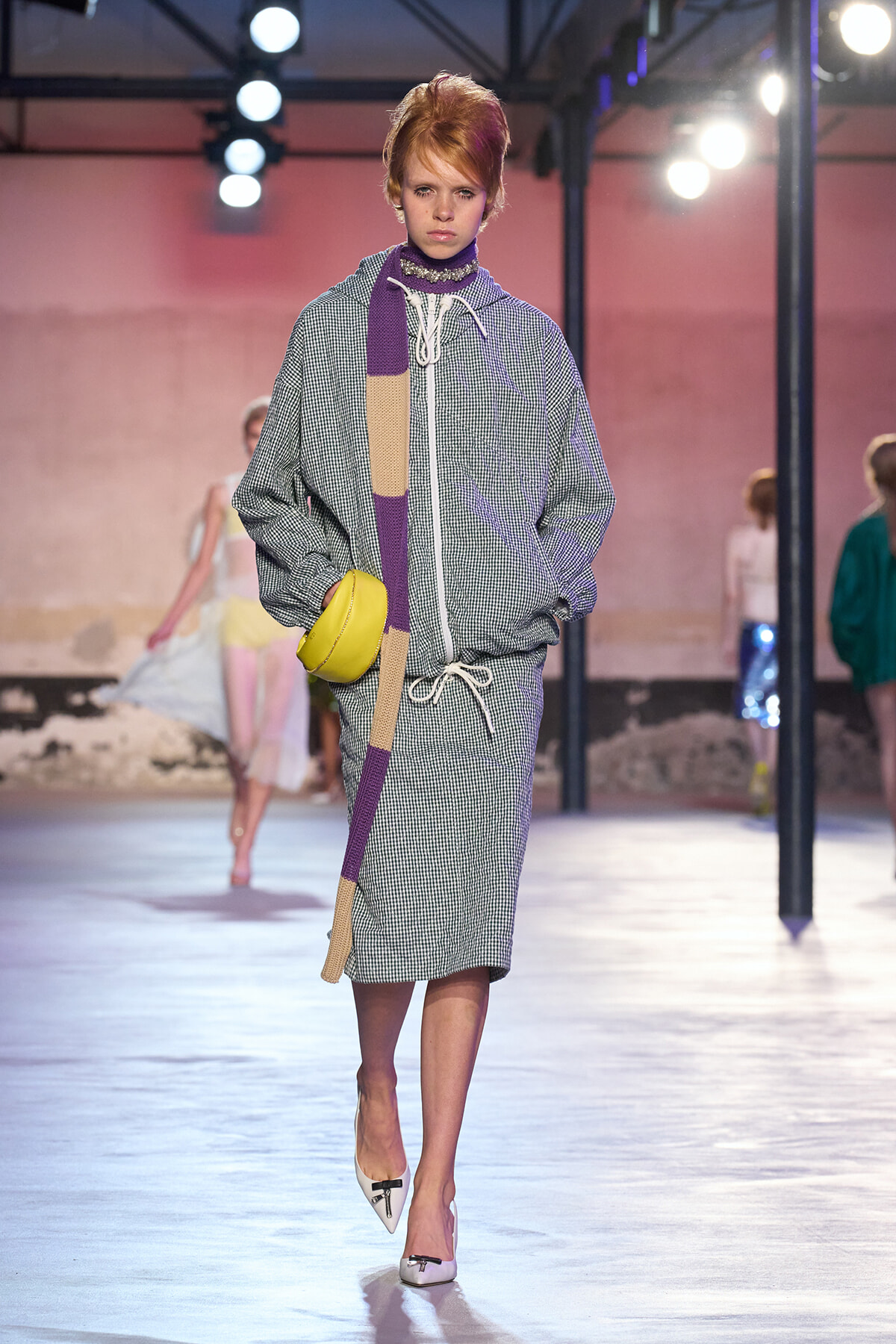 Model walking a runway wearing a gray checkered hooded jumpsuit with a long purple-beige scarf and a yellow crossbody bag, confident stride.