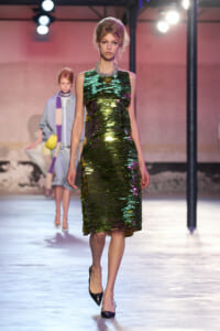 Model on a runway wearing a sleeveless, knee-length green metallic sequin dress with a chunky necklace and black heels.