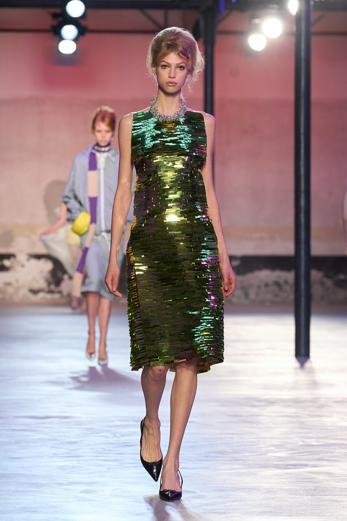 Model on a runway wearing a sleeveless, knee-length green metallic sequin dress with a chunky necklace and black heels.