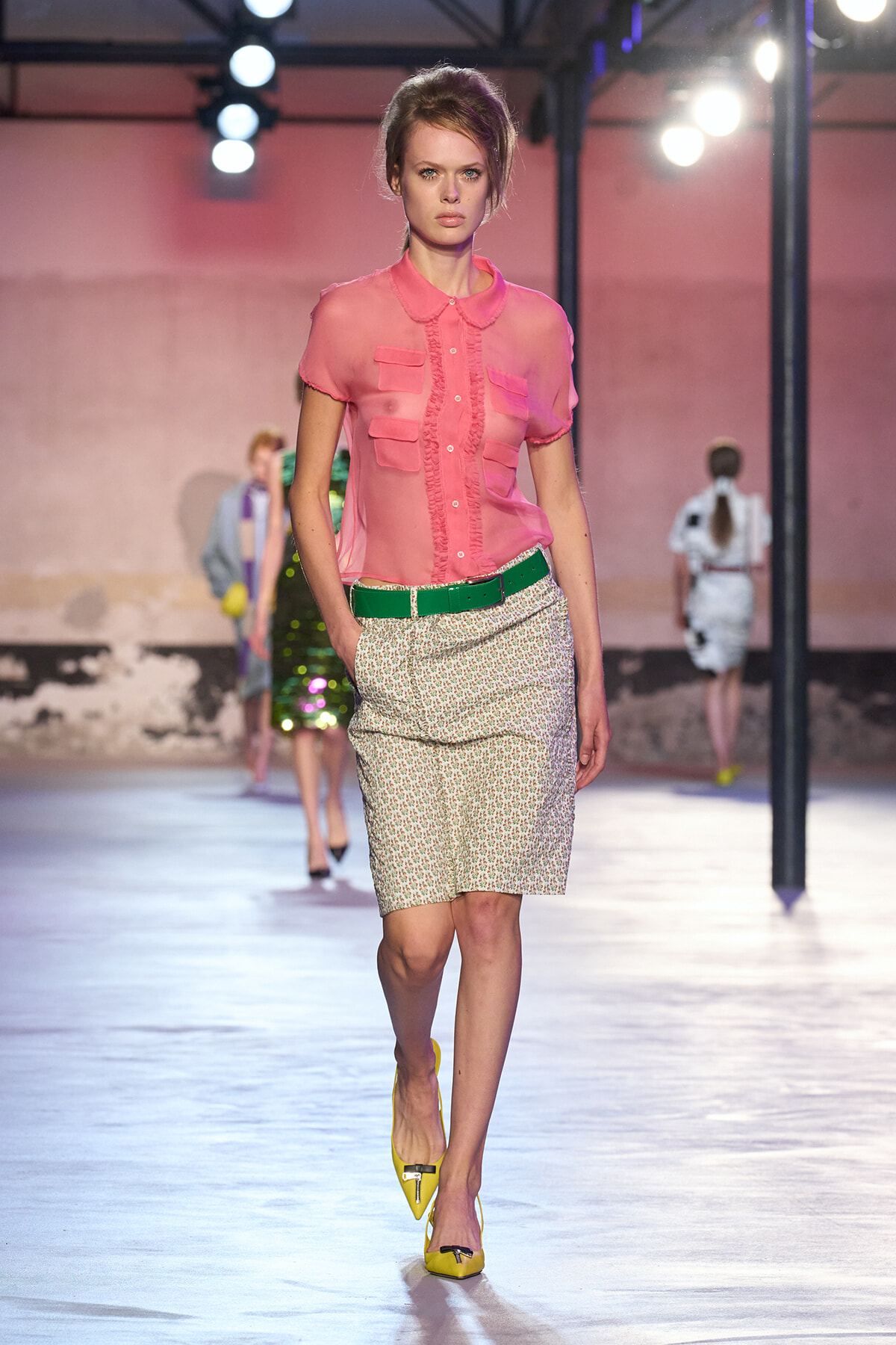 Model walking a runway in a pink sheer blouse with ruffle trim and pockets, a green belt, a beige textured pencil skirt, and yellow pointed-toe heels.