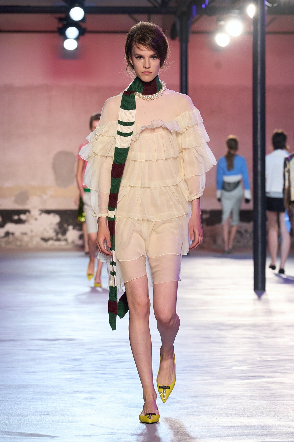 Model walks the runway in a cream, multi-tiered off-shoulder dress with a pearl necklace, paired with a long green striped scarf and yellow flats on a fashion show catwalk.