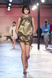Model walks a metallic gold brocade mini dress on a runway, hands in pockets, under bright stage lights.