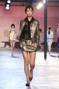 Female model walks the runway in a metallic gold brocade blazer and matching mini skirt, black blouse, and pointed heels.
