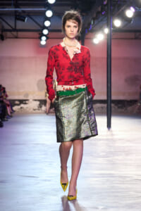 Model walking a fashion runway in a red floral cardigan, pearl necklace, metallic skirt, green belt, and yellow heels.