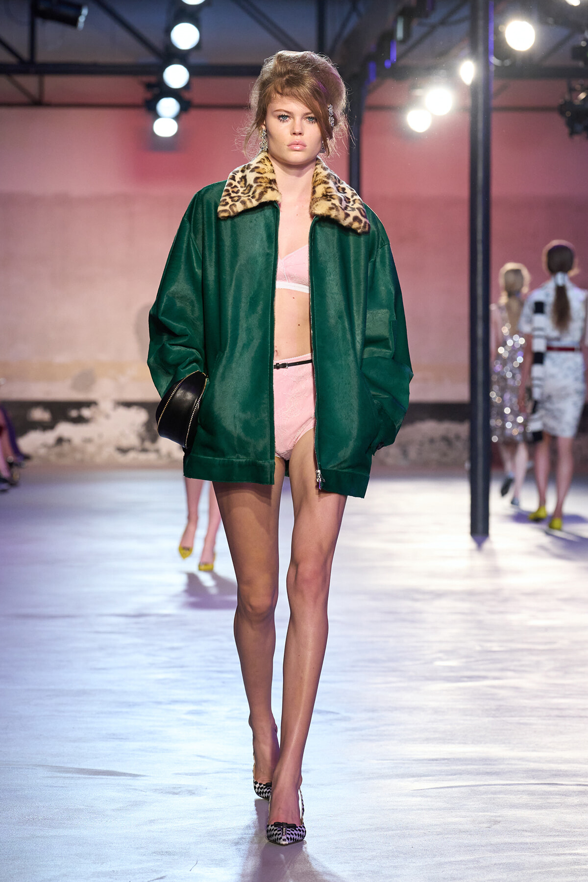 Model on a fashion runway wearing a green oversized jacket with a leopard-print collar over pink lingerie, walking toward the camera.