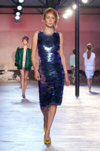 Model walks the runway in a knee-length blue sequin dress with a chunky silver necklace under bright stage lights.