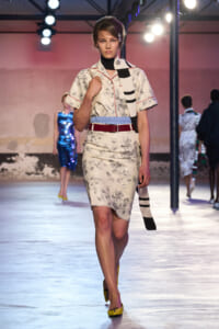 Model walks the runway in a cream floral dress with red belt and a black-and-white scarf over the shoulder, yellow shoes visible