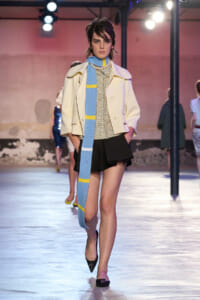 Model walking a fashion runway in a cream oversized coat, floral blouse, black shorts, and a light blue scarf with yellow stripes.