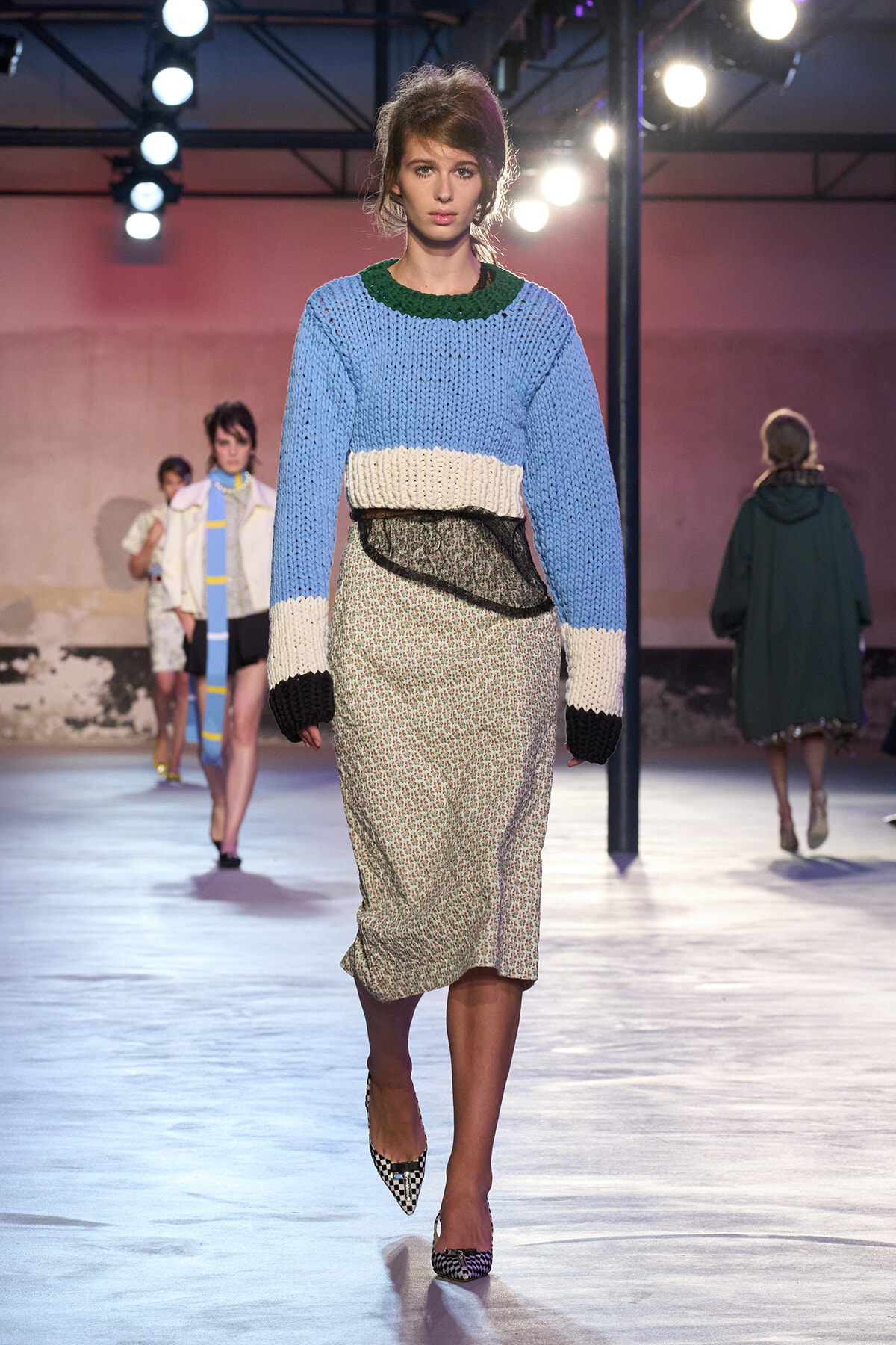 Model walking a fashion runway in a blue chunky knit sweater with cream cuffs and hem, over a floral midi skirt, wearing black-and-white checkered heels.