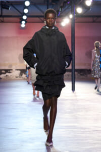 Model walks the runway in an oversized black hooded jacket with a layered black mini skirt, white shoes, under bright stage lights.