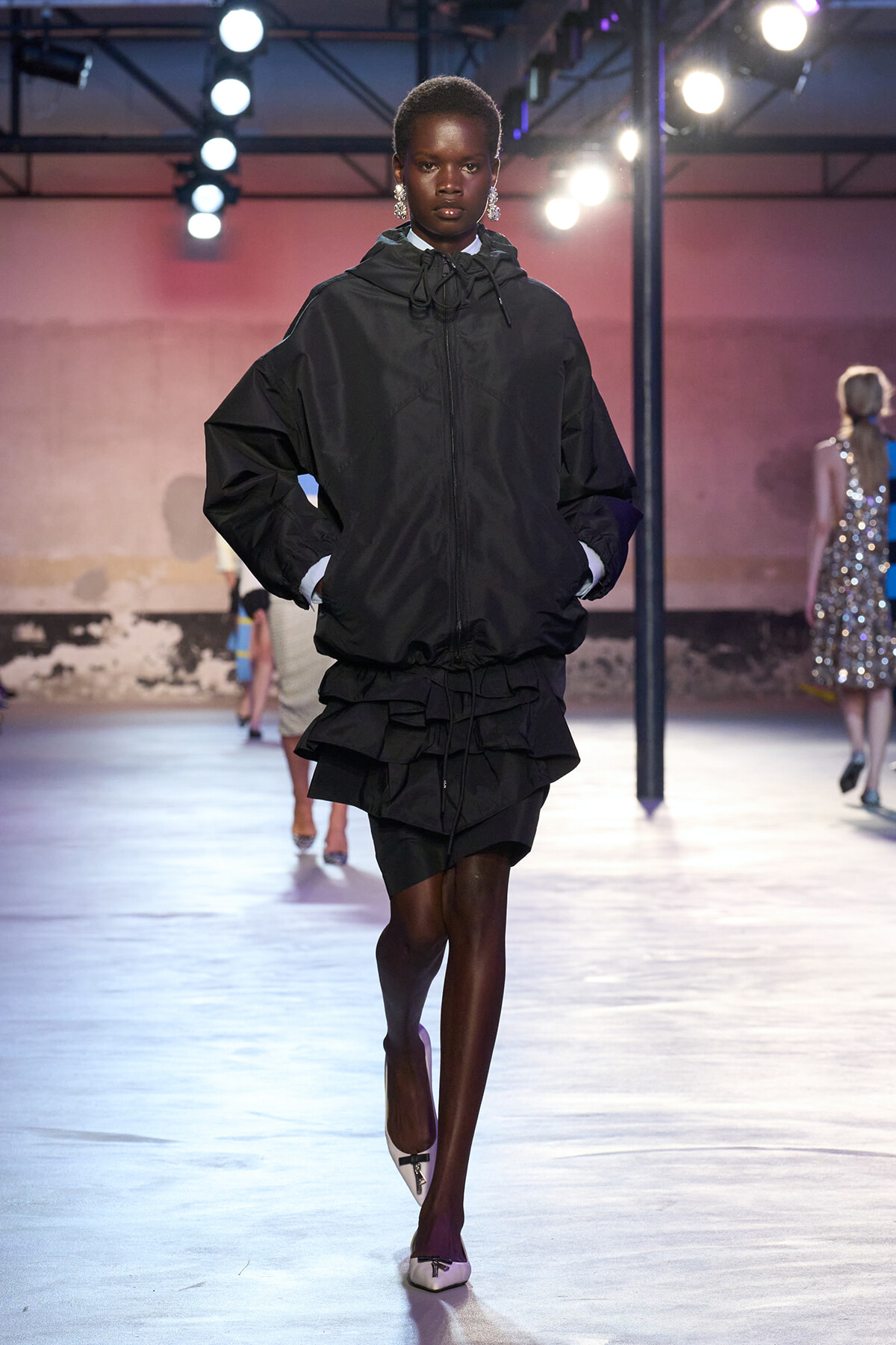 Model walks the runway in an oversized black hooded jacket with a layered black mini skirt, white shoes, under bright stage lights.