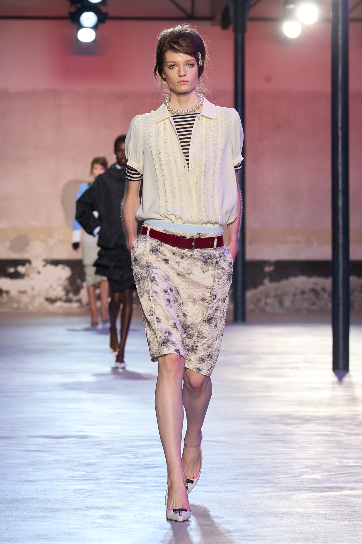 Model walking a fashion runway in an ivory blouse, striped top, floral knee-length skirt, and red belt; pearl necklace and white flats.