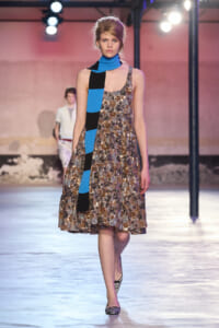 Model walking a fashion runway in a knee-length sequined dress with a long blue-and-black scarf draped over her shoulder and neck.