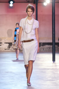 Model walking a fashion runway in a white blouse, white skirt, brown belt, and layered pearl necklaces.