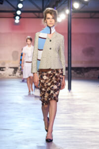 Model walks a fashion runway wearing a light floral blazer, brown belt, and a shimmering floral pencil skirt.