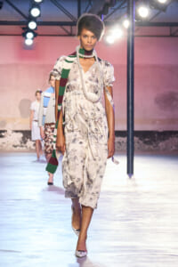 Model walking a fashion runway in a light floral dress with a long pearl necklace and a green-and-white striped scarf draped over her shoulder under bright stage lights.