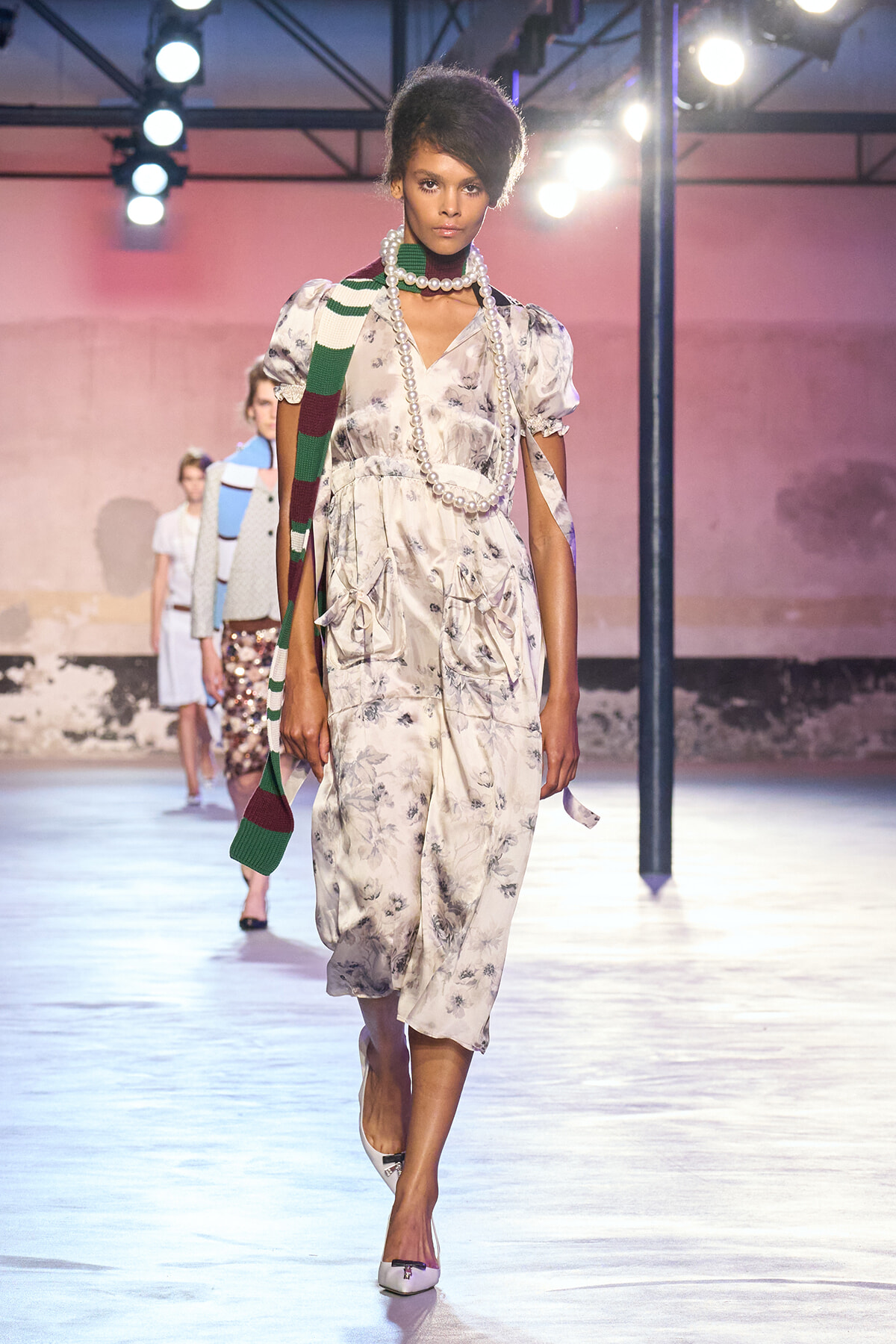 Model walking a fashion runway in a light floral dress with a long pearl necklace and a green-and-white striped scarf draped over her shoulder under bright stage lights.