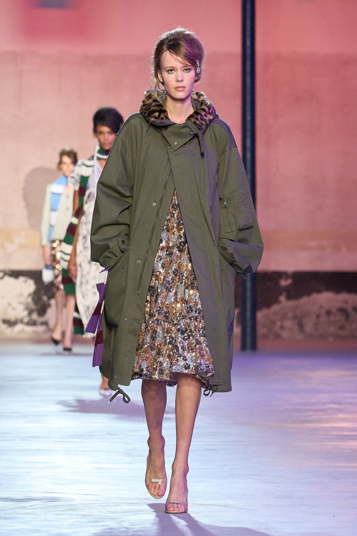 Model walks the runway in an oversized olive parka with a fur-trimmed hood over a shimmering gold sequin dress, pink backdrop behind her.