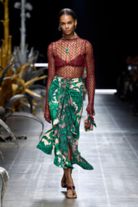 Model walking a runway in a burgundy fishnet top over a matching bra, a green tropical-print wrap skirt, and a green pendant necklace; carrying a small clutch with flowers.