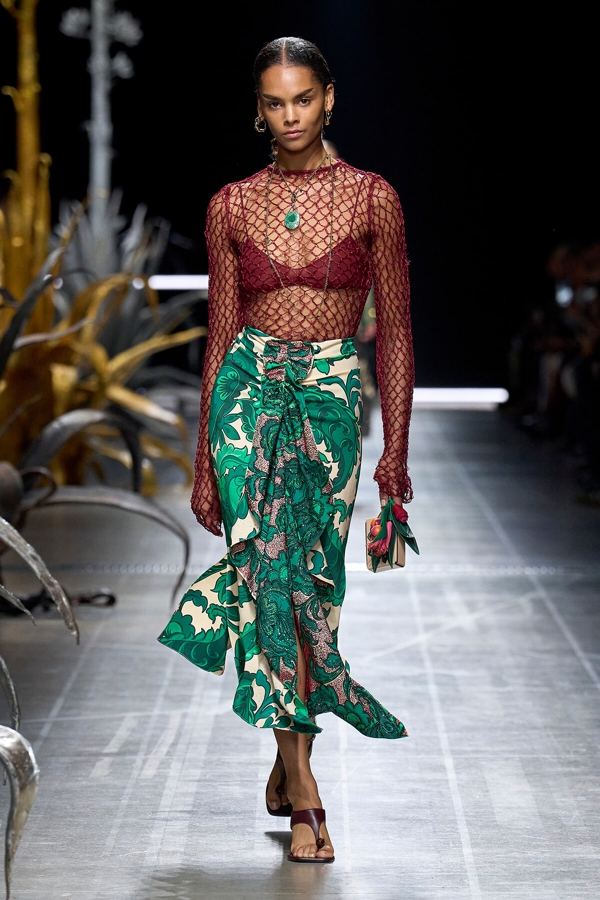 Model walking a runway in a burgundy fishnet top over a matching bra, a green tropical-print wrap skirt, and a green pendant necklace; carrying a small clutch with flowers.