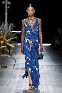 Model walks a runway in a blue floral print dress with sheer black mesh long sleeves, carrying a small green-and-blue handbag and wearing sandals.